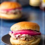 A pulled pork sandwich with coleslaw and pickled onions on a black plate, with another sandwich and condiments blurred in the background. Text reads “Pulled Pork Sandwiches.”.