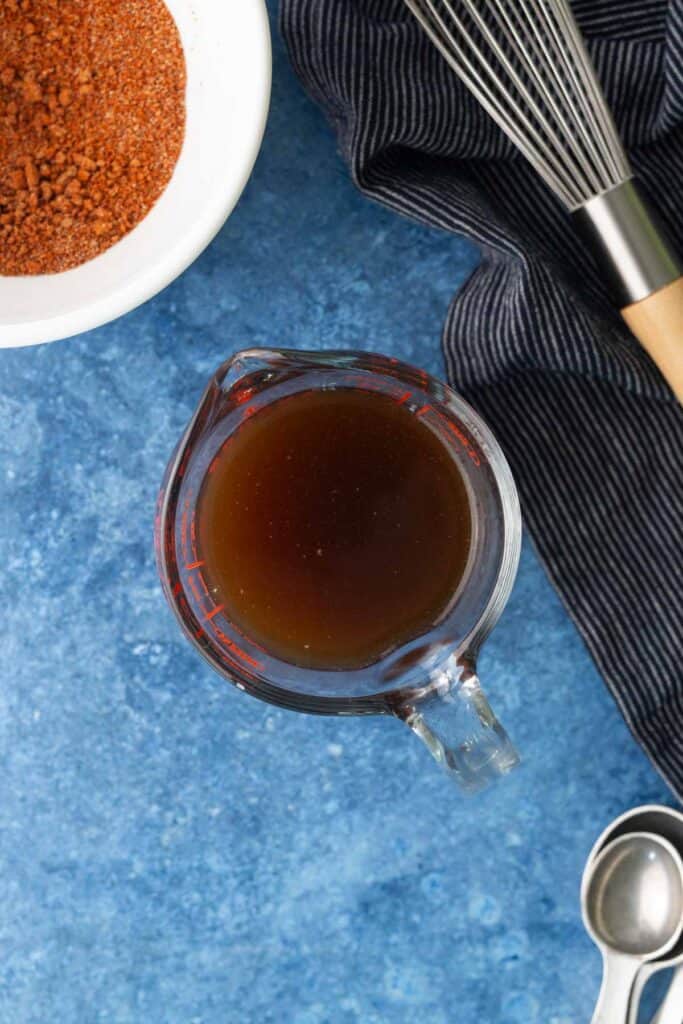 A glass measuring cup filled with brown liquid sits on a blue surface next to a bowl of spices, a whisk, a striped cloth, and a metal measuring spoon.