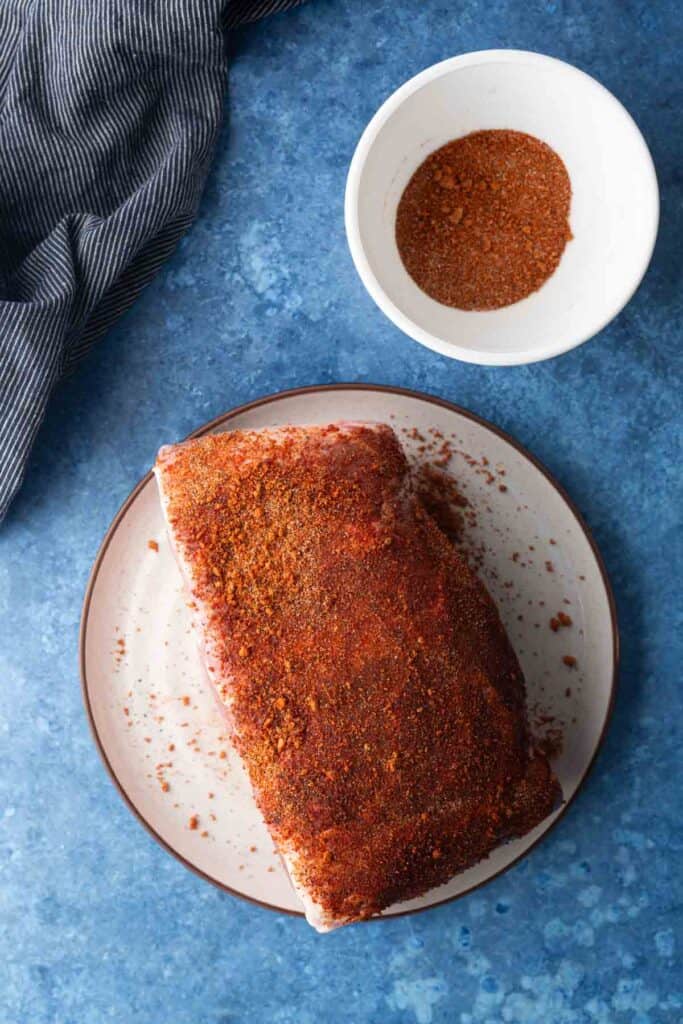 A slab of raw meat covered in dry spice rub sits on a plate next to a bowl containing extra seasoning, on a blue surface.