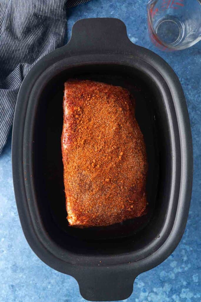 A seasoned pork roast sits in a black slow cooker on a blue surface, with a striped cloth and an empty measuring cup nearby.