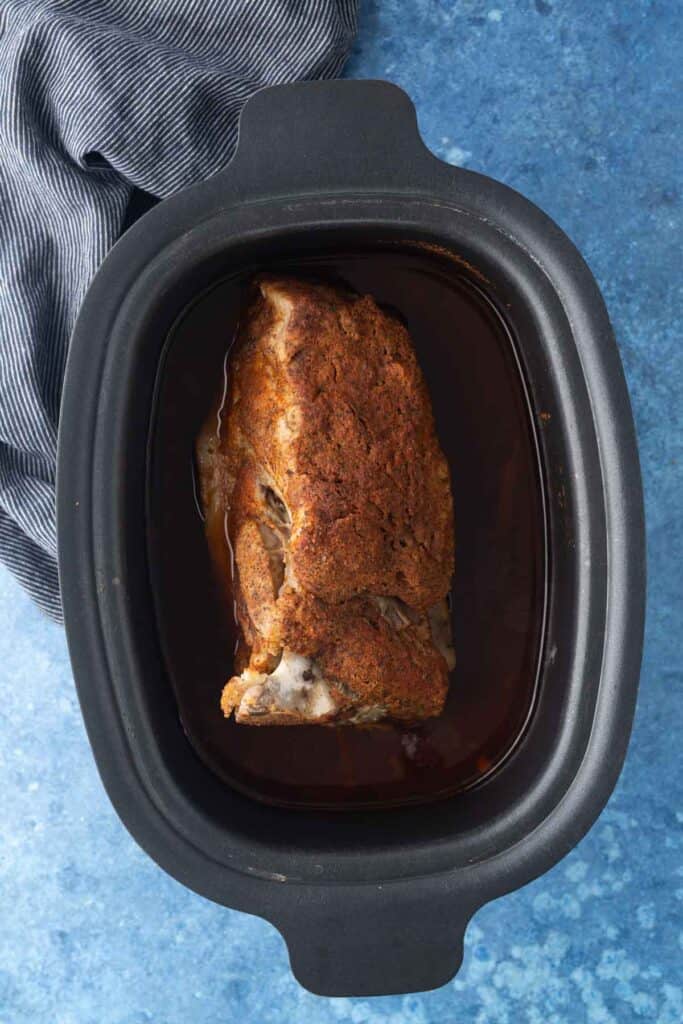 A seasoned pork roast sits in dark cooking liquid inside a black slow cooker on a blue surface next to a striped cloth.