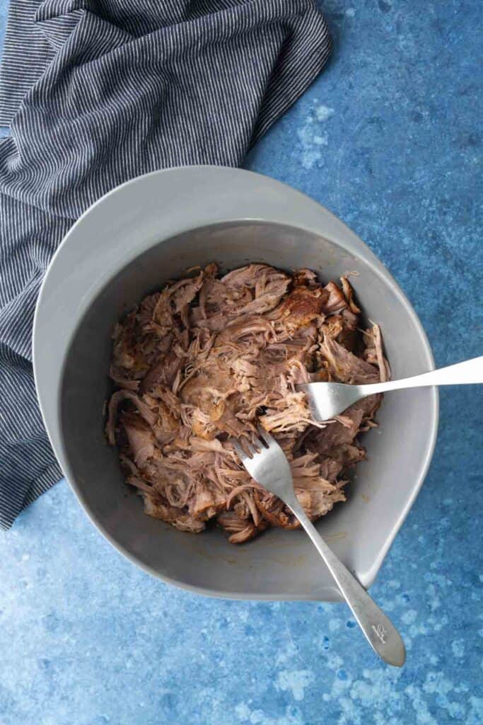 Shredded cooked meat in a gray mixing bowl with two forks, on a blue surface next to a striped cloth.