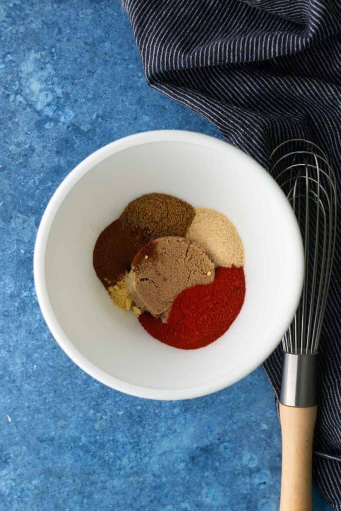 A white bowl containing piles of various ground spices and brown sugar sits on a blue countertop next to a black-and-white striped cloth and a whisk.