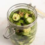 A glass jar filled with sliced cucumbers, dill, mustard seeds, and peppercorns in brine on a white surface.