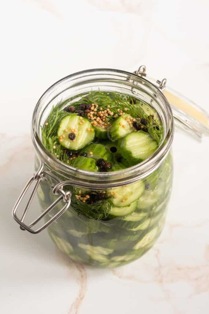 Refrigerator Dill Pickles A glass jar filled with sliced cucumbers, dill, mustard seeds, and peppercorns in brine on a white surface.