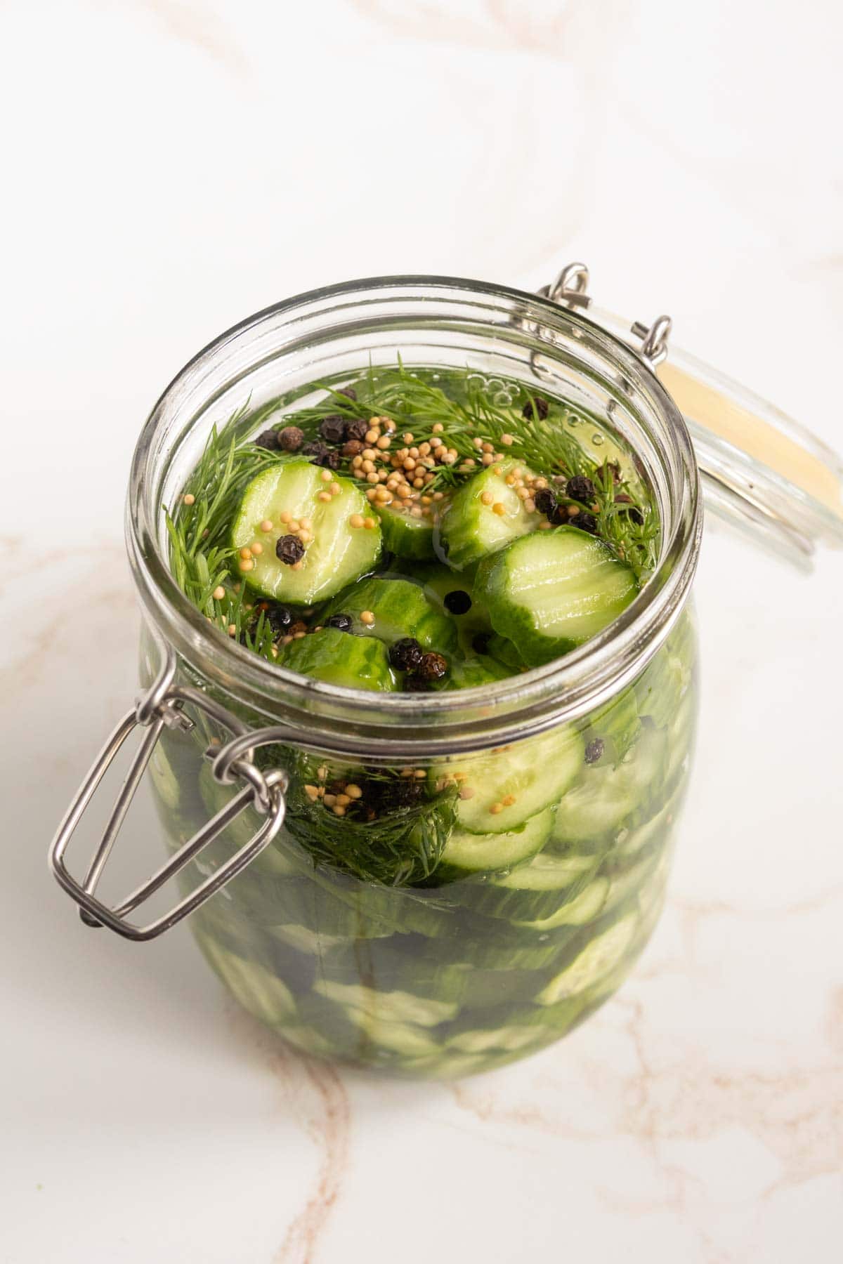 A glass jar filled with sliced cucumbers, dill, mustard seeds, and peppercorns in brine on a white surface.