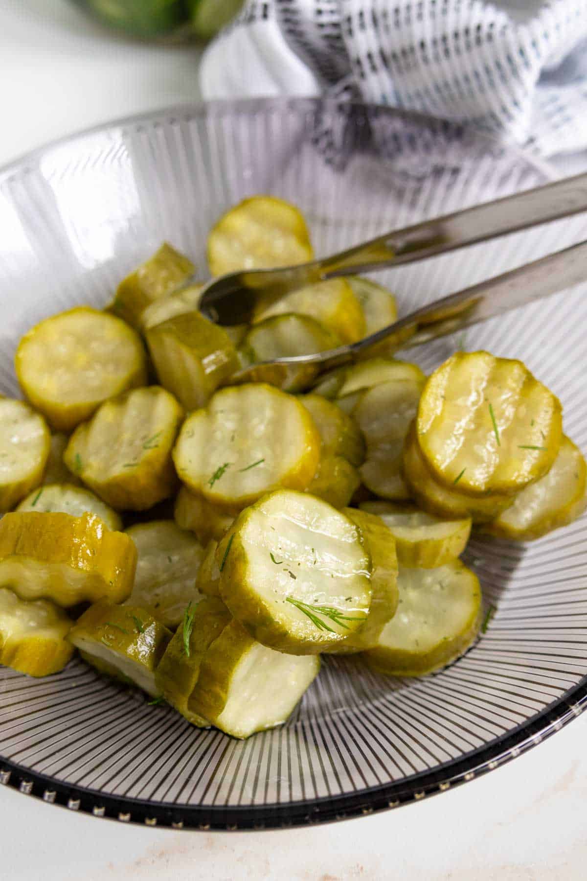 A glass plate with sliced pickles and metal tongs resting on top, with a striped cloth in the background.