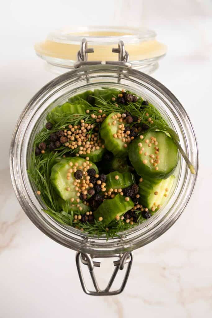 A glass jar filled with sliced cucumbers, dill, mustard seeds, and black peppercorns, prepared for pickling on a white surface.