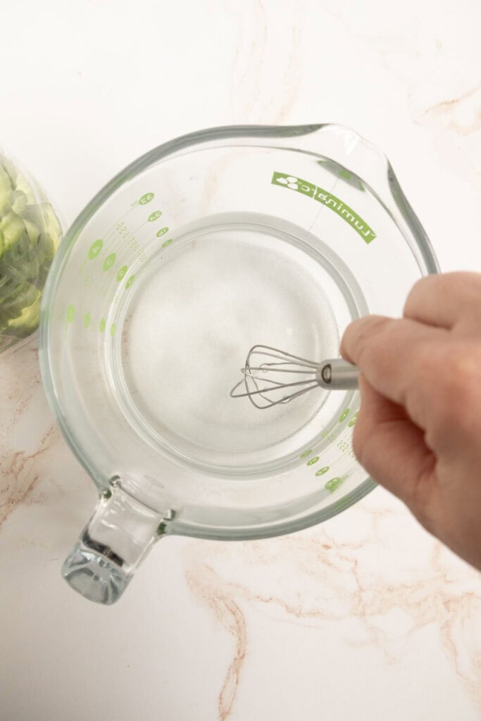 A hand whisking sugar into water in a glass measuring cup on a light-colored countertop.