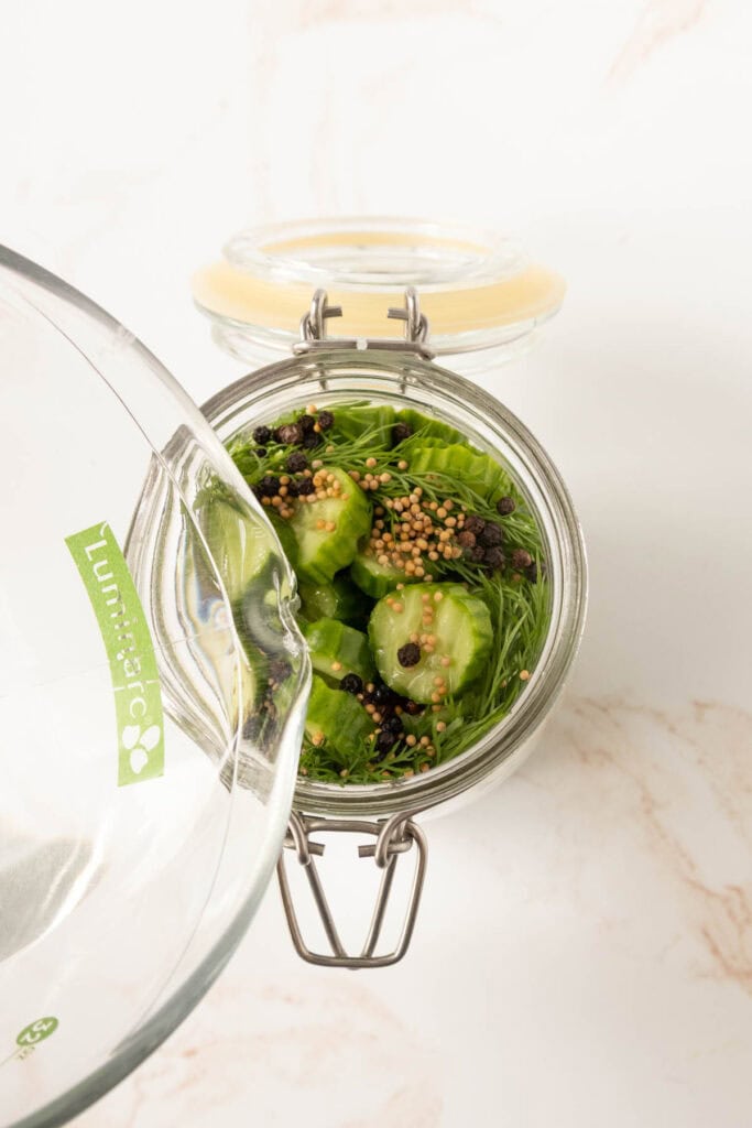 A glass jar filled with sliced cucumbers, dill, mustard seeds, and peppercorns is being filled with liquid from a glass measuring cup.