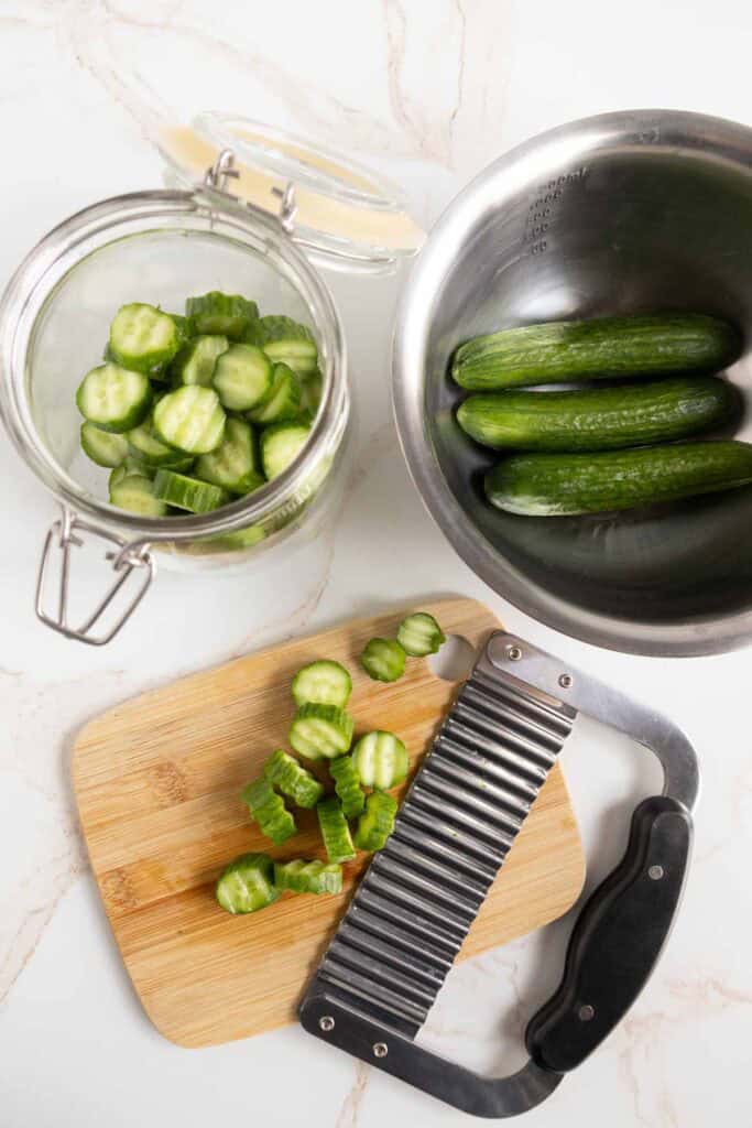 Sliced cucumbers on a wooden board with a crinkle cutter, whole cucumbers in a metal bowl, and sliced cucumbers in a glass jar on a white surface.
