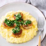 Seared scallops topped with green herb sauce served on a bed of creamy yellow polenta, arranged on a white plate with a fork on the side.
