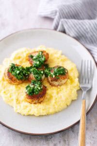 Seared scallops topped with green herb sauce served on a bed of creamy yellow polenta, arranged on a white plate with a fork on the side.
