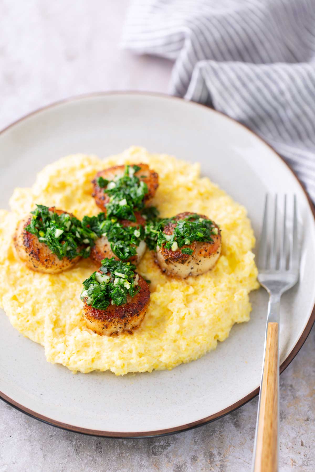Seared scallops topped with green herb sauce served on a bed of creamy yellow polenta, arranged on a white plate with a fork on the side.