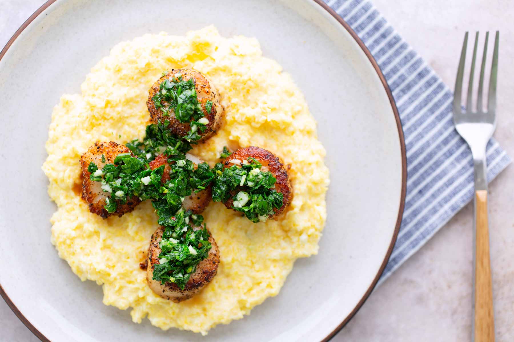 Four seared scallops with green herb topping served on a bed of creamy yellow polenta, on a white plate next to a blue-striped napkin and a fork.