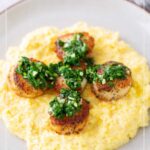 Five seared scallops topped with green herb garnish are served on a bed of creamy yellow polenta on a plate.