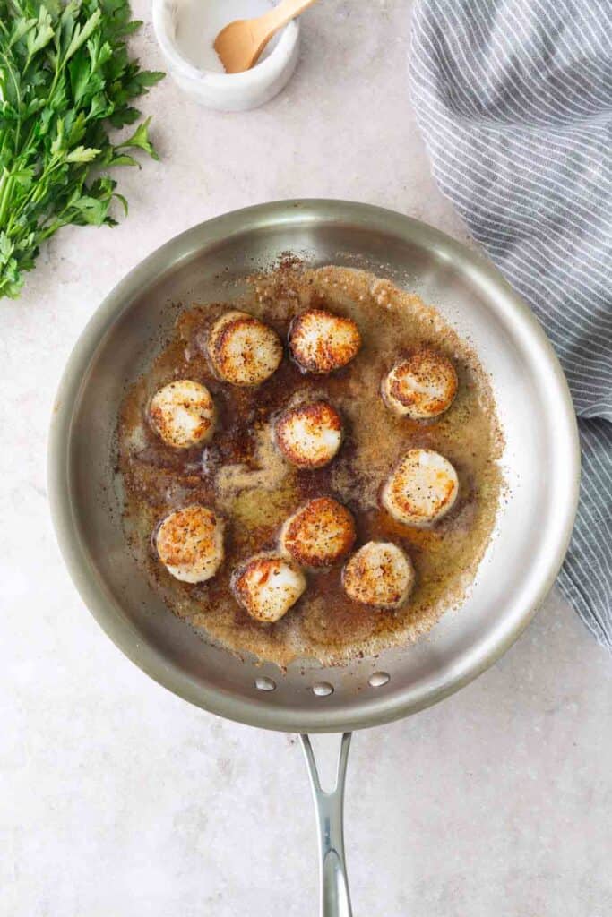 Seared scallops cooking in a stainless steel pan with browned butter, next to a bunch of parsley and a striped kitchen towel.