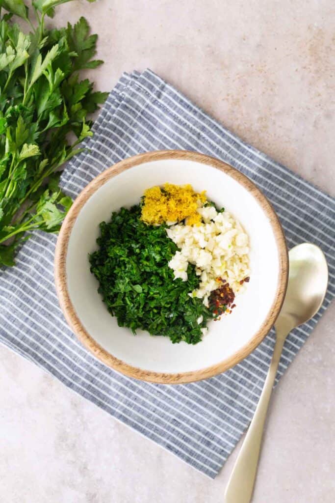 A bowl containing chopped parsley, minced garlic, lemon zest, and red pepper flakes sits on a gray striped cloth next to a spoon and fresh parsley.