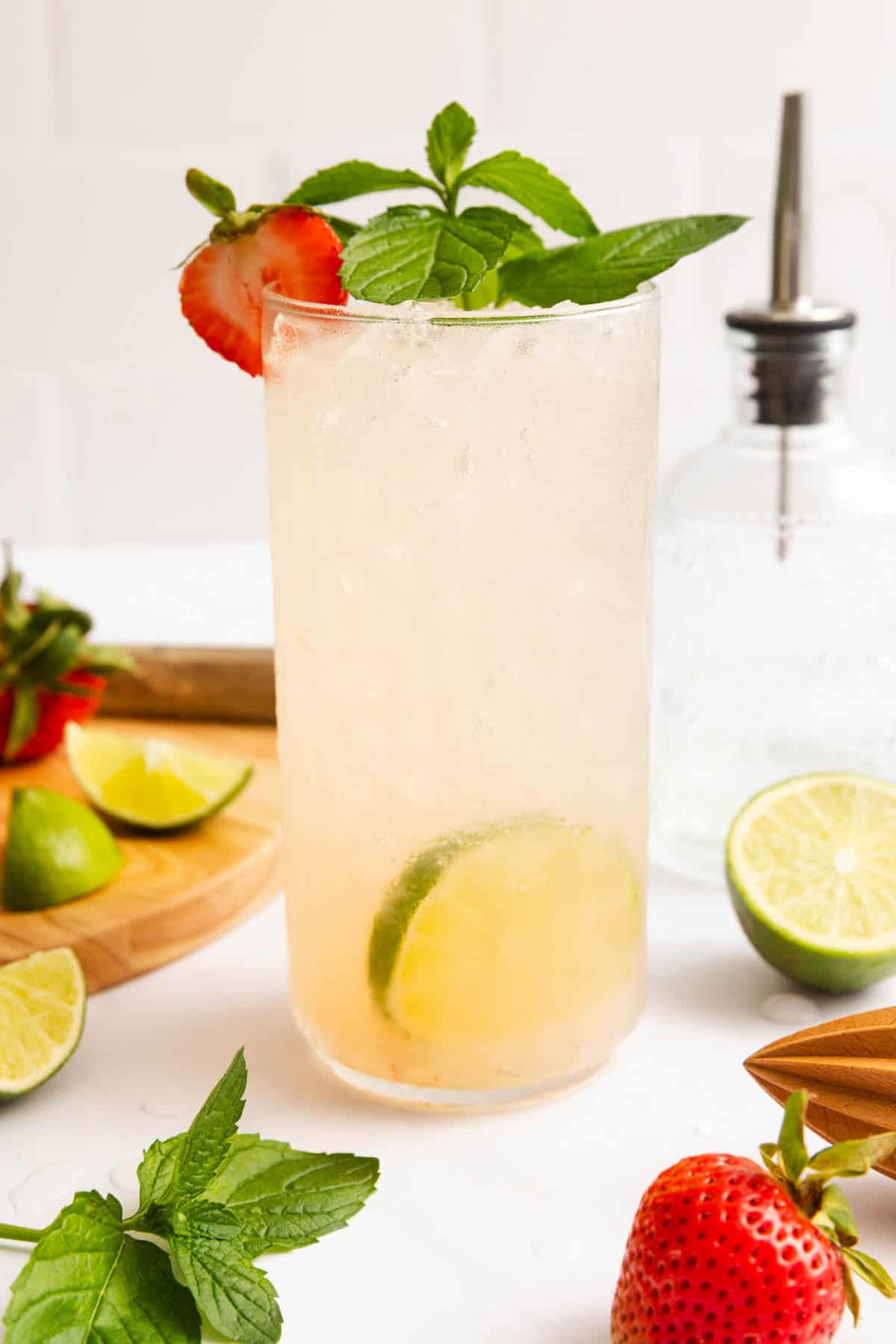 A tall glass of iced drink garnished with a lime slice, strawberry, and mint, placed on a white surface with fresh limes, strawberries, and mint nearby.