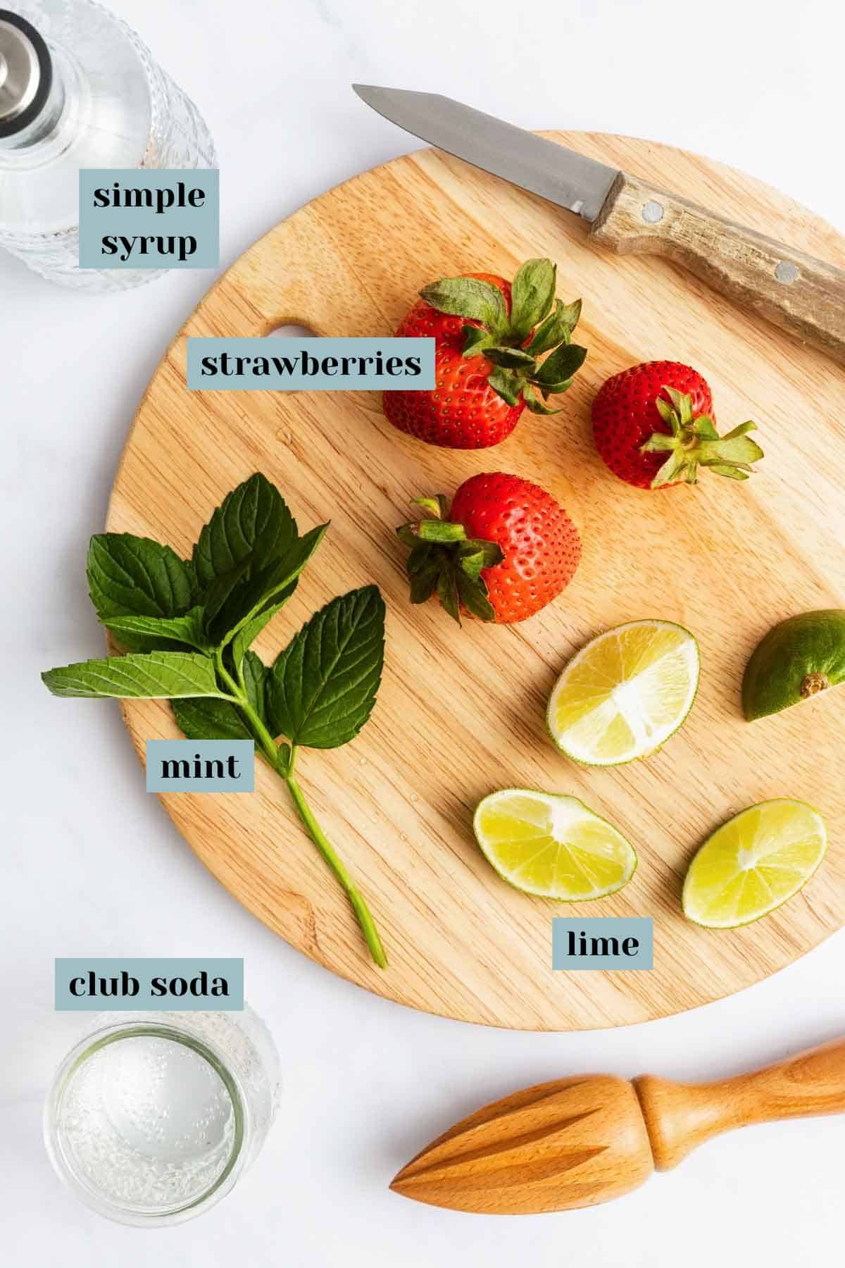A wooden cutting board with strawberries, lime wedges, and mint, surrounded by a knife, wooden citrus juicer, bottle of club soda, and glass of simple syrup.