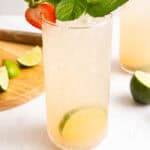 A tall glass with a light-colored drink, garnished with a strawberry, mint leaves, and lime slices. Lime wedges and a strawberry are on the table nearby.