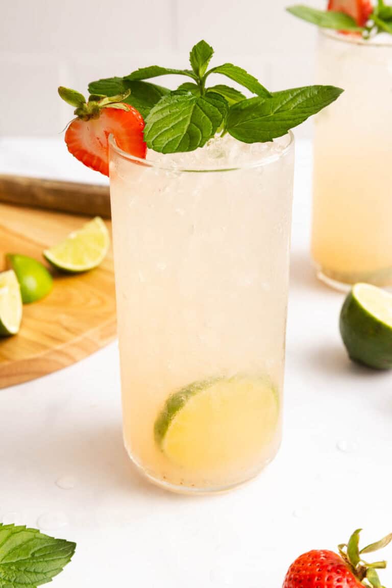 A tall glass with a light-colored drink, garnished with a strawberry, mint leaves, and lime slices. Lime wedges and a strawberry are on the table nearby.