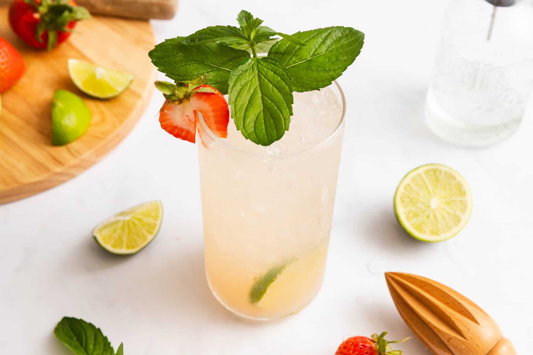 A tall glass of iced cocktail garnished with mint leaves and a strawberry, surrounded by lime wedges, strawberries, and a wooden citrus juicer on a white surface.