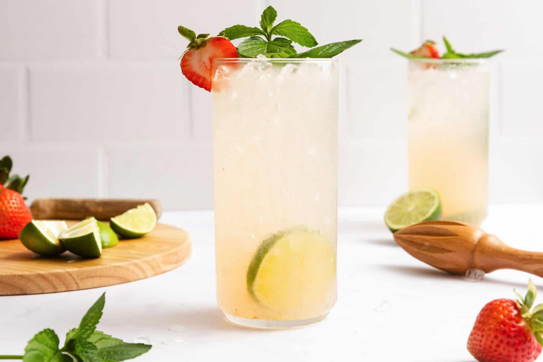 A tall glass of iced lemonade garnished with a strawberry slice and mint leaves, with lime wedges inside. Another similar glass, fresh strawberries, and lime wedges are in the background.
