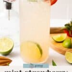 A tall glass of mint strawberry mocktail garnished with mint and a strawberry slice, with lime wedges and a drink shaker in the background.