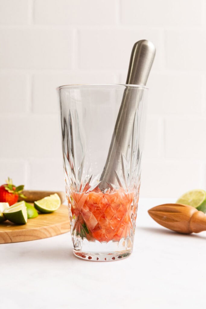 A metal muddler in a crystal glass with muddled strawberries and limes; lime wedges and strawberries on a wooden board in the background.