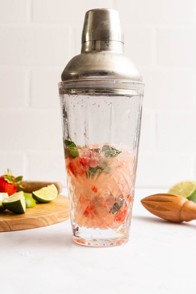 A cocktail shaker filled with muddled strawberries, lime, and mint sits on a counter beside a wooden board with strawberries and lime wedges.