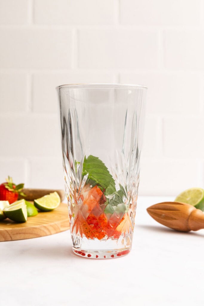 A clear glass with mint leaves and muddled strawberries at the bottom, with lime wedges, a strawberry, and a wooden citrus reamer on a cutting board in the background.
