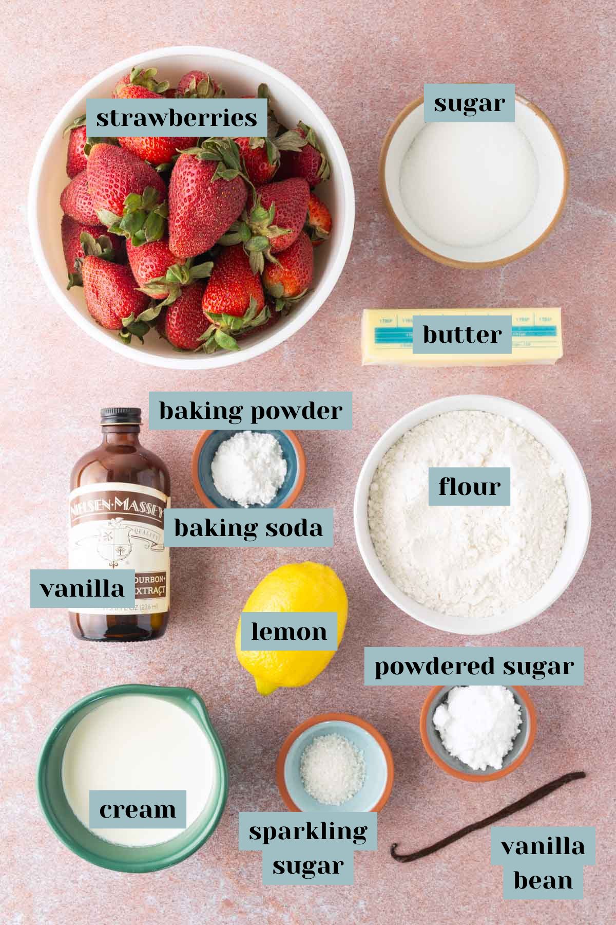 Ingredients for strawberry shortcake laid out on a countertop, including strawberries, sugar, butter, flour, cream, lemon, vanilla, baking powder, baking soda, powdered sugar, and vanilla bean.