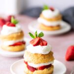 A plate with a strawberry shortcake topped with whipped cream, sliced strawberries, and a mint leaf, with more shortcakes and strawberries in the background.