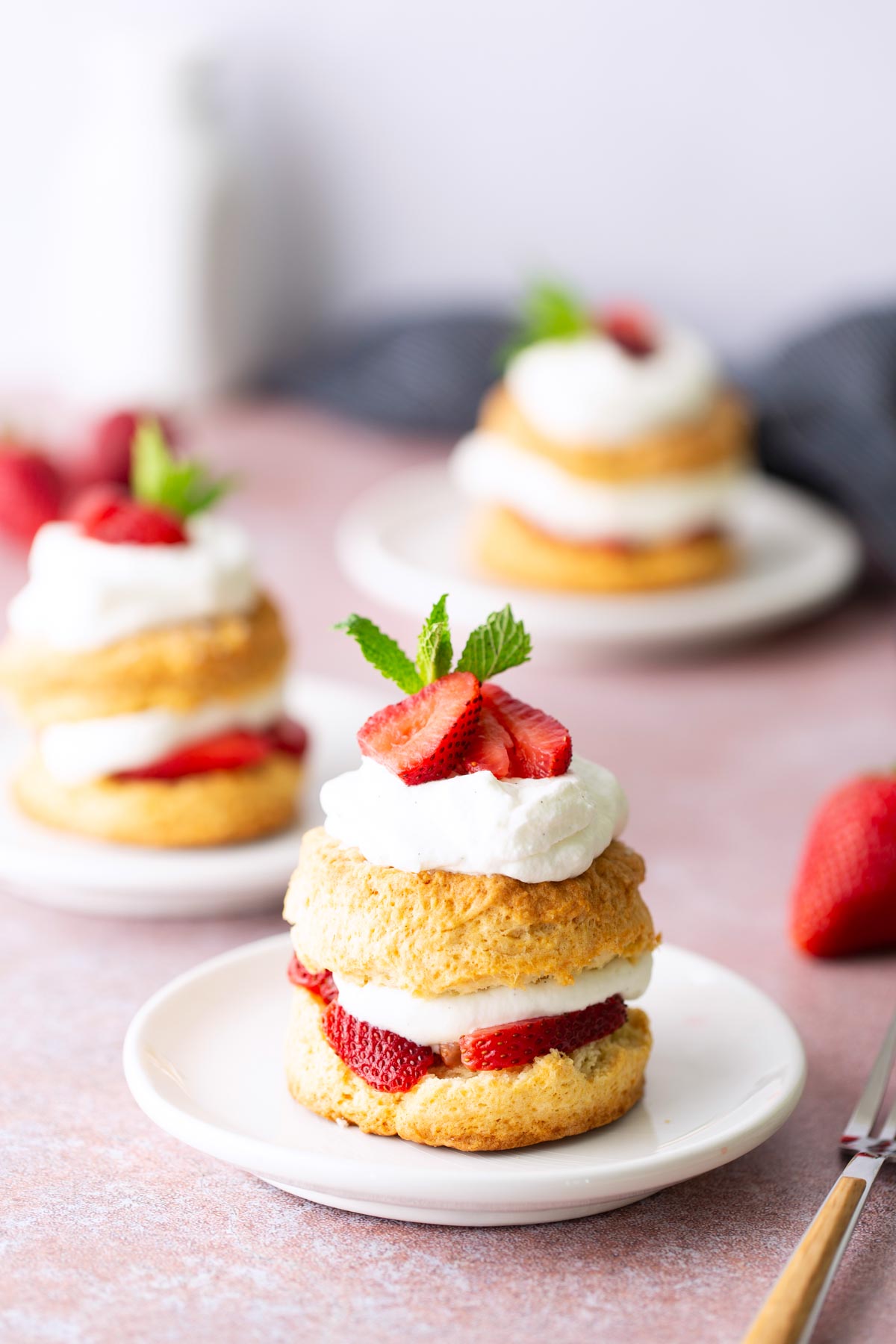 A plate with a strawberry shortcake topped with whipped cream, sliced strawberries, and a mint leaf, with more shortcakes and strawberries in the background.