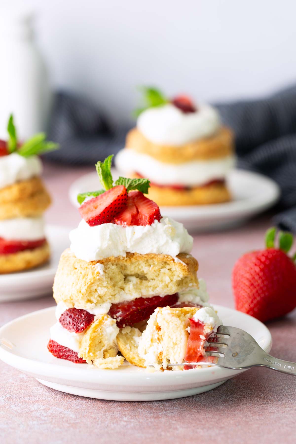 Three plates of strawberry shortcake with layers of biscuit, whipped cream, and sliced strawberries; a fork rests on the front plate with a piece cut out.
