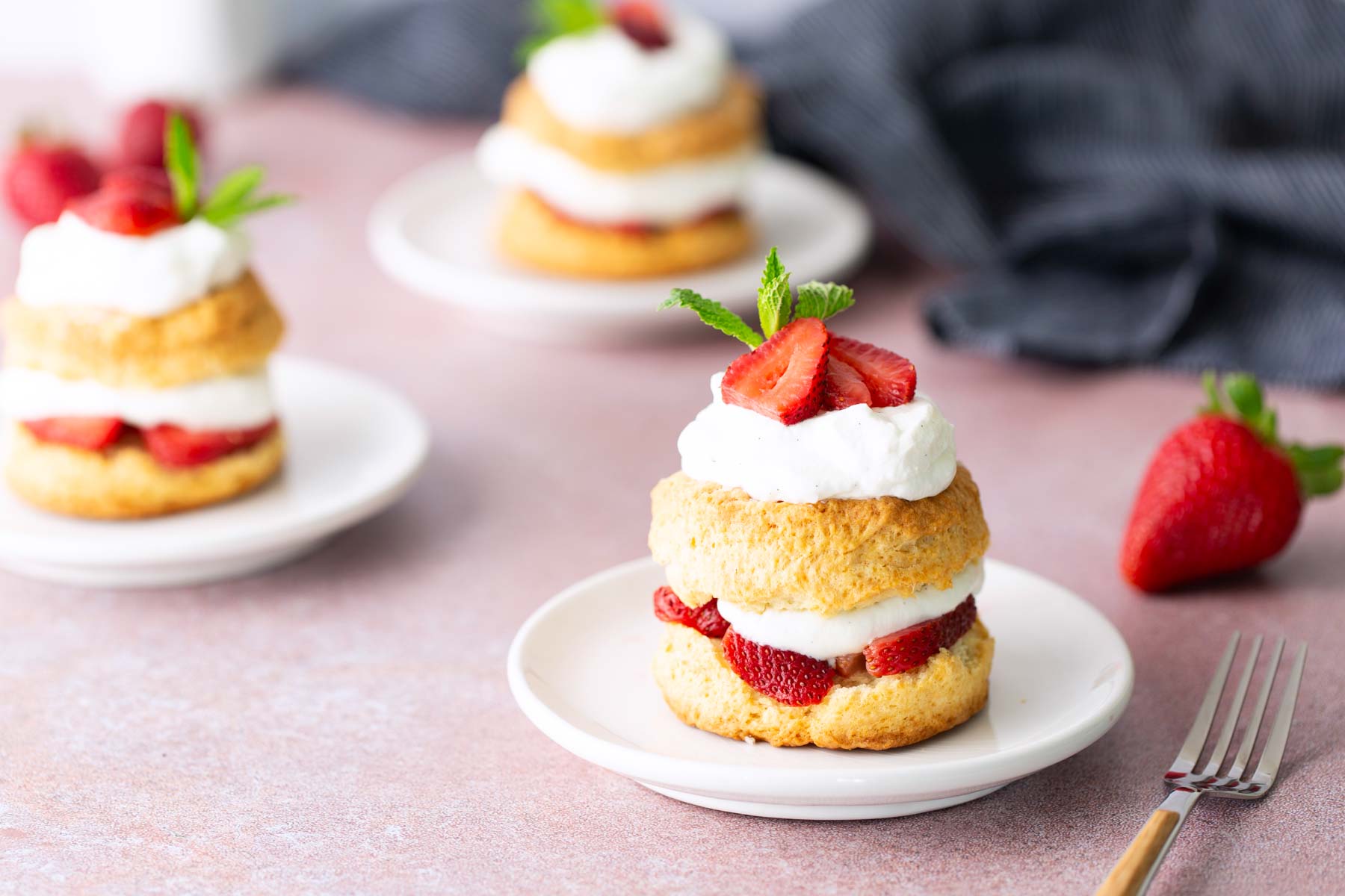 Three strawberry shortcakes with whipped cream are served on white plates, garnished with strawberry slices and mint, with a fork and whole strawberry nearby.