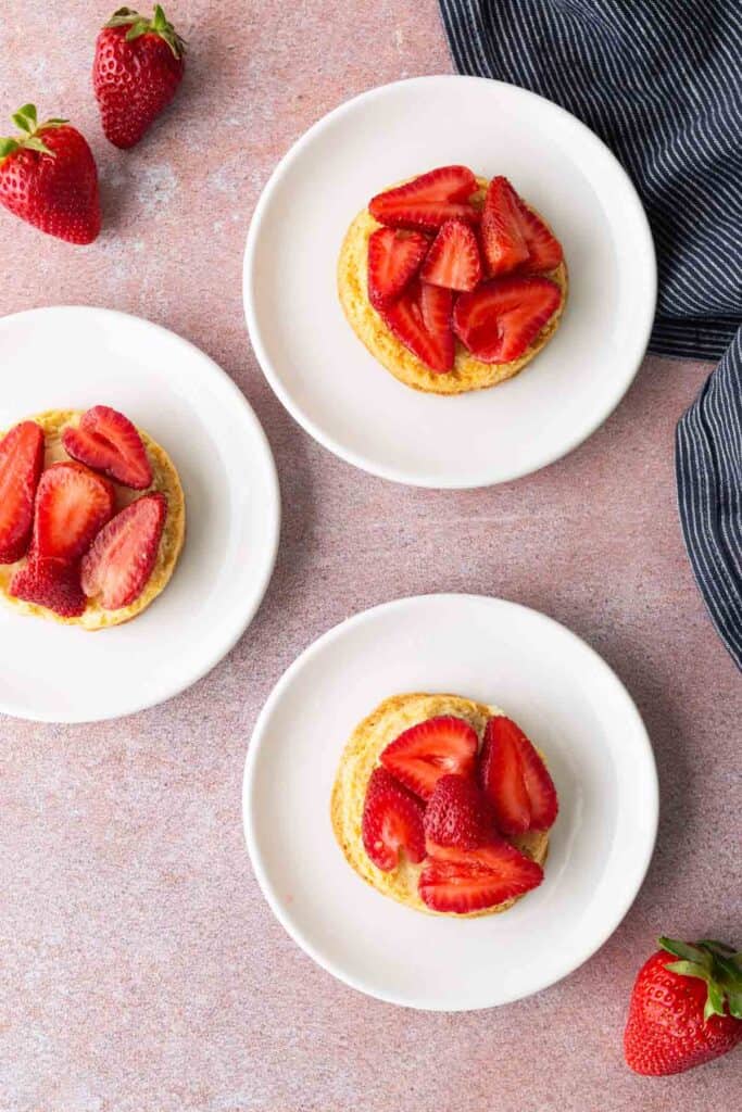 Three white plates each with a biscuit topped with sliced strawberries, placed on a pink surface with whole strawberries and a striped cloth nearby.