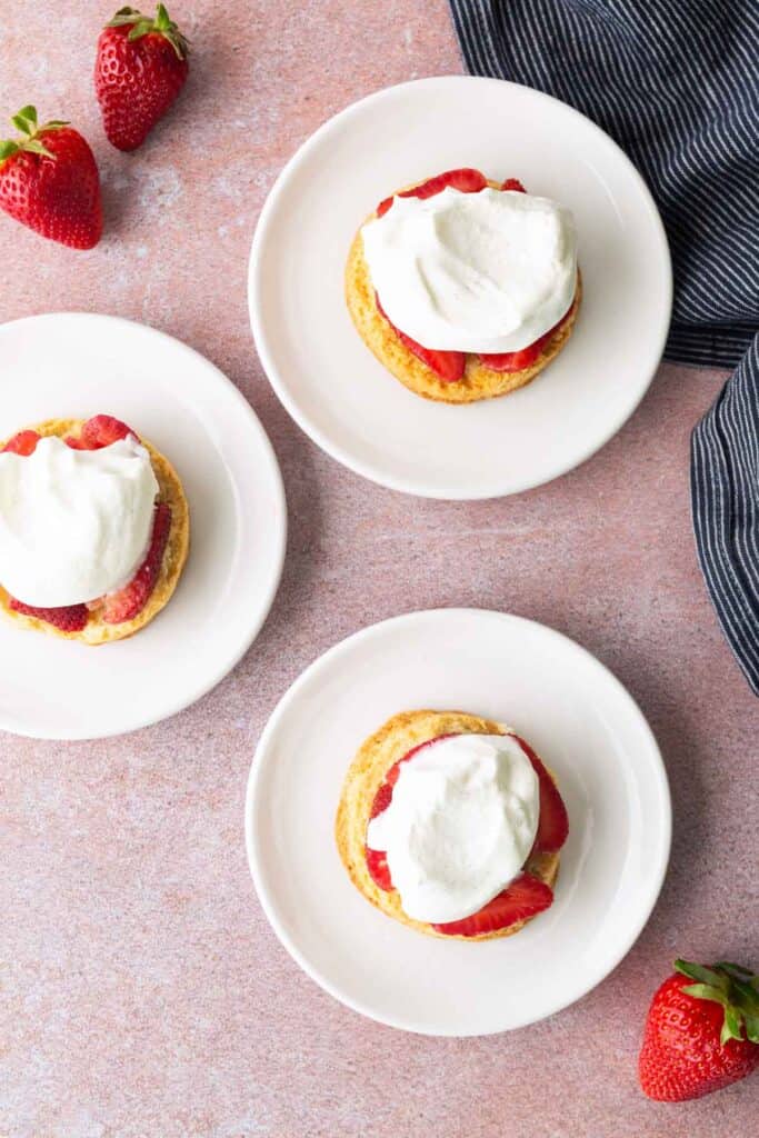 Three plates with strawberry shortcakes topped with whipped cream, surrounded by fresh strawberries, on a pink surface with a striped napkin.