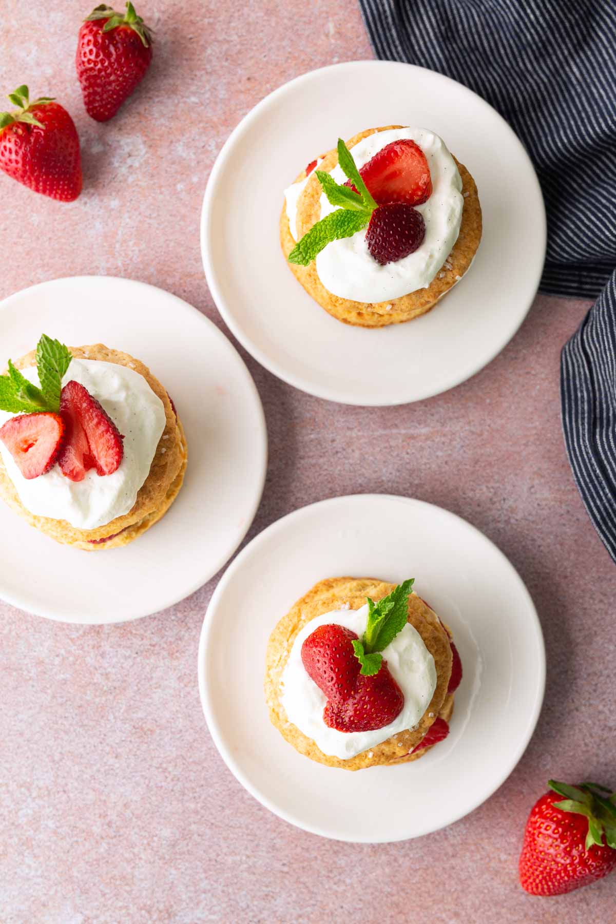 Three strawberry shortcakes topped with whipped cream, fresh strawberries, and mint leaves are arranged on white plates, with whole strawberries and a striped cloth nearby on a pink surface.