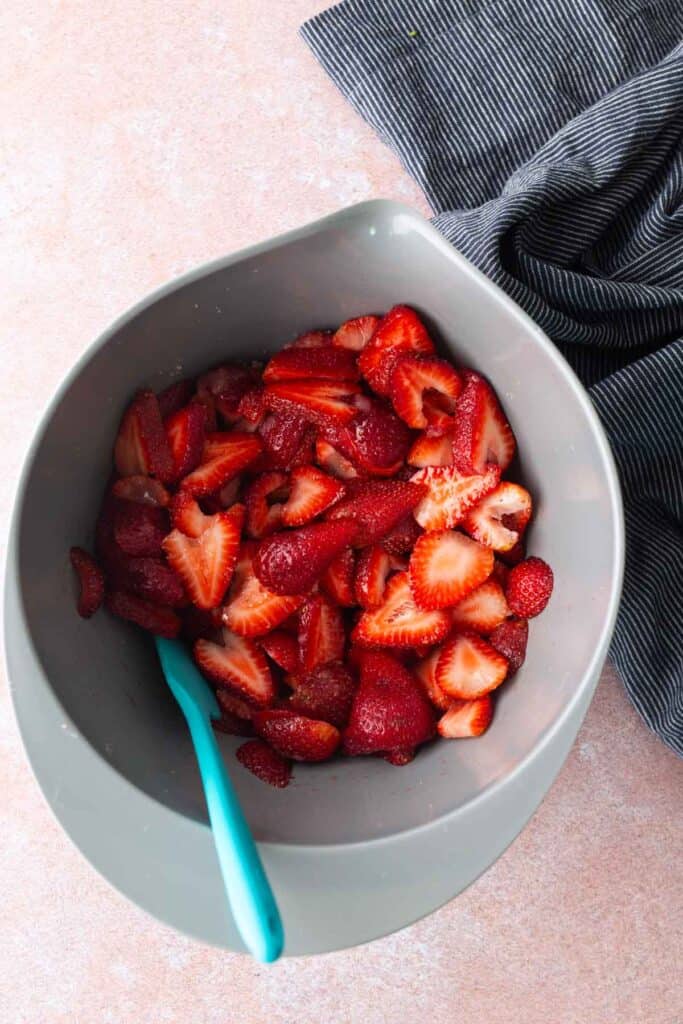 A gray bowl filled with sliced strawberries and a blue spoon, placed on a light surface next to a dark striped cloth.