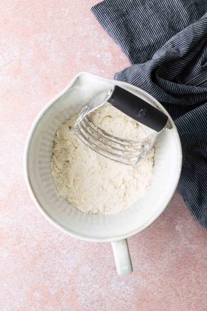 A pastry cutter resting in a bowl filled with flour mixture, placed on a pink surface next to a dark striped cloth.