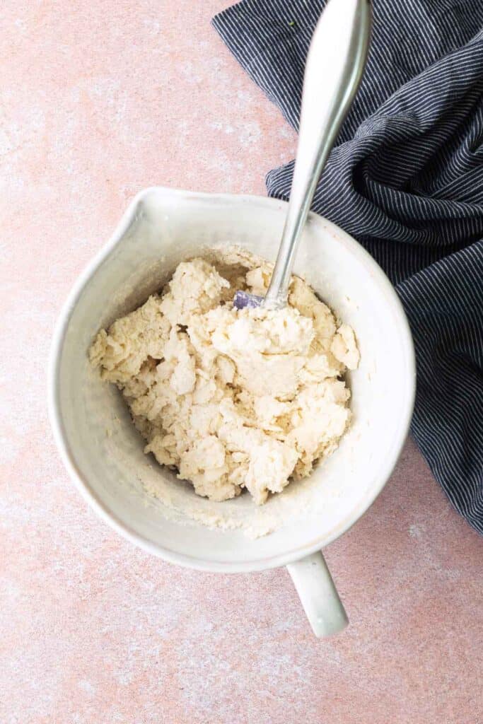 A white mixing bowl with crumbly dough and a metal spoon inside, placed on a pink surface next to a dark striped cloth.