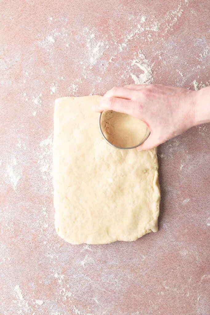 A hand uses a round metal cutter to cut dough on a floured surface.