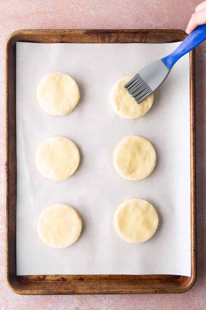 A baking tray lined with parchment paper holds six round biscuit dough pieces. A hand applies an egg wash with a brush to the top right biscuit.