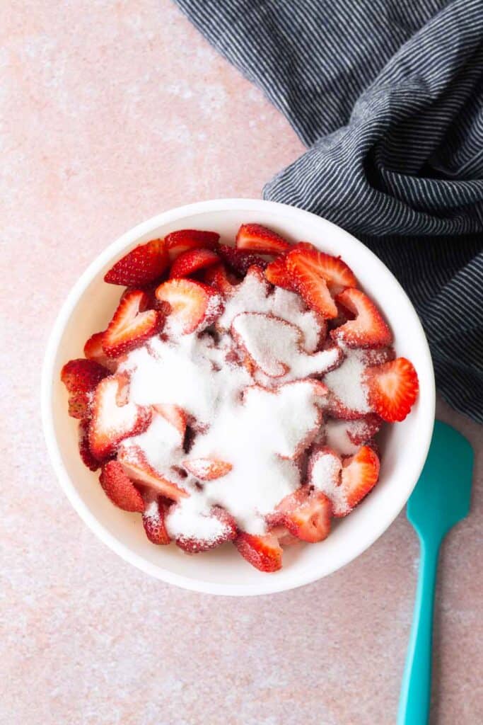Sliced strawberries in a white bowl topped with granulated sugar, next to a blue spatula and a dark striped cloth on a light pink surface.