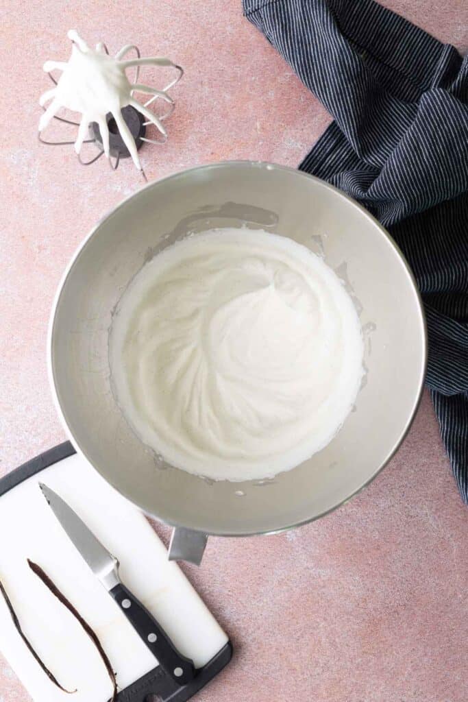 A metal mixing bowl with whipped egg whites, a whisk with more egg whites, a cutting board with a knife, and a scraped vanilla bean pod next to a dark cloth.