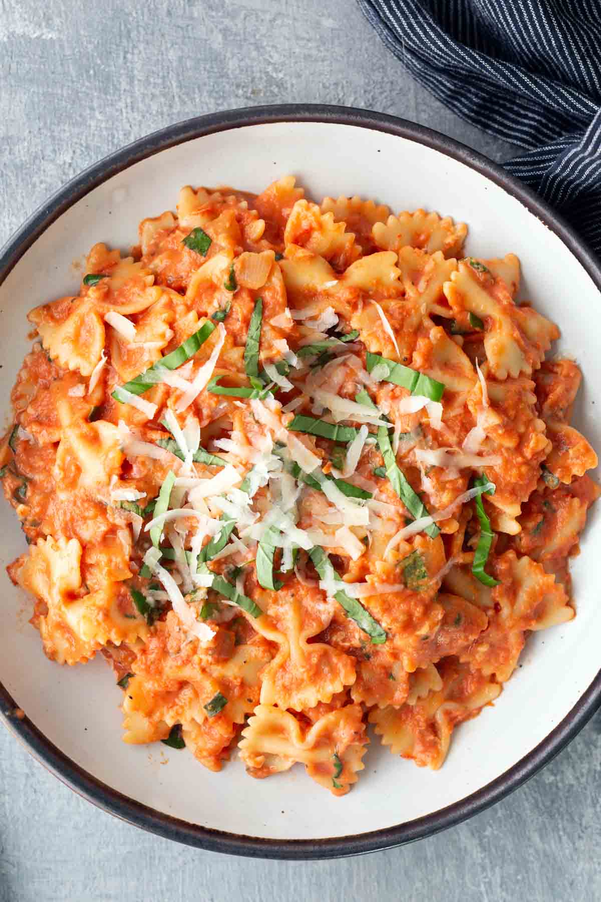 A bowl of bow tie pasta in tomato sauce, topped with grated cheese and fresh basil, on a light surface with a striped cloth nearby.