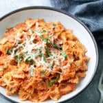 A bowl of farfalle pasta in a creamy tomato sauce, topped with shredded cheese and fresh basil, sits on a gray surface next to a fork and a dark striped cloth.