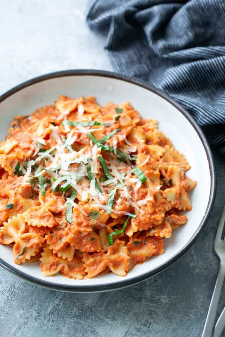 A bowl of farfalle pasta in a creamy tomato sauce, topped with shredded cheese and fresh basil, sits on a gray surface next to a fork and a dark striped cloth.